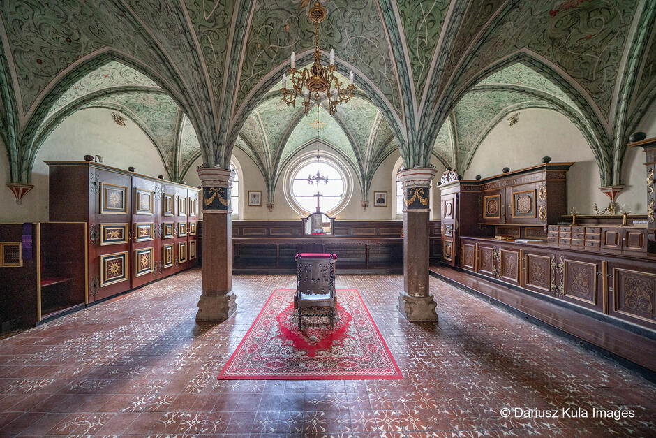 Zdjęcie przedstawia piękne, zabytkowe wnętrze o gotyckiej architekturze. Znajduje się w nim bogato zdobiony sufit z zielonymi sklepieniami pokrytymi misternymi wzorami roślinnymi. Całość wspierają eleganckie filary ozdobione złoconymi detalami. Centralnym punktem pomieszczenia jest dekoracyjne krzesło ustawione na czerwonym, wzorzystym dywanie, co nadaje przestrzeni dostojnego i ceremonialnego charakteru. W tle, na osi kompozycji, znajduje się okrągłe okno wpuszczające światło, pod którym widnieje ołtarzyk z krzyżem. Po bokach pomieszczenia ustawione są drewniane szafy i zdobione szafki o bogatej ornamentyce, wskazujące na historyczne i sakralne przeznaczenie wnętrza. Posadzka jest udekorowana geometrycznymi wzorami, które harmonijnie komponują się z całością wystroju.