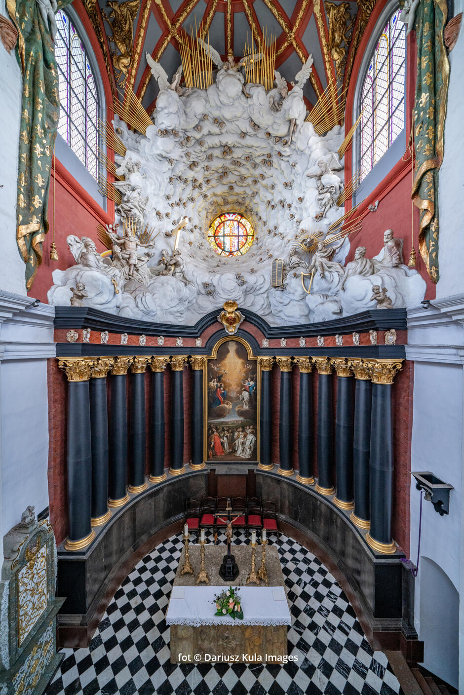 Zdjęcie przedstawia niezwykle bogato zdobione wnętrze barokowej kaplicy lub ołtarza, pełne dynamiki i teatralnego efektu charakterystycznego dla tego stylu. Centralnym punktem jest ołtarz z malowidłem religijnym, otoczony monumentalnymi, czarnymi kolumnami ze złotymi kapitelami, które nadają całości dostojny i majestatyczny wygląd. Najbardziej imponującym elementem kompozycji jest iluzjonistyczna dekoracja nad ołtarzem, przedstawiająca niebiańską scenę. Białe, rzeźbione chmury wypełniają przestrzeń, a wśród nich widać liczne anioły i postaci świętych. Całość sprawia wrażenie, jakby sklepienie świątyni otwierało się na niebo, co potęguje efekt mistycyzmu. W centrum znajduje się okrągłe, witrażowe okno, przez które wpada światło, rozjaśniając scenę i dodając jej boskiego blasku.