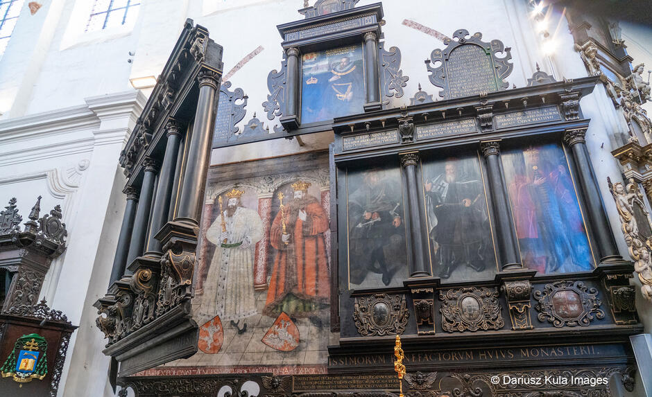Zdjęcie przedstawia imponującą ścienną kompozycję nagrobno-memorialną, prawdopodobnie związaną z ważnymi postaciami historycznymi lub fundatorami klasztoru czy kościoła. Struktura łączy malowidła, rzeźbione elementy oraz ciemne, bogato zdobione drewniane obramowania, które nadają całości monumentalny charakter. Centralnym elementem jest duże malowidło przedstawiające dwie postaci w ceremonialnych strojach – jedna w białych szatach, druga w czerwonych. Obie trzymają insygnia władzy, co może sugerować, że są to królowie lub dostojnicy kościelni. Obok, w czarnych obramowaniach, znajdują się portrety innych ważnych osób, prawdopodobnie związanych z fundacją tego miejsca. Powyżej widnieją inskrypcje w języku łacińskim, upamiętniające ich zasługi.