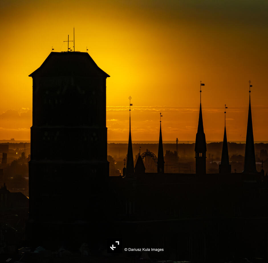 Zdjęcie ukazuje majestatyczną sylwetkę gotyckiej świątyni na tle złocistego zachodu słońca. Mroczne kontury masywnej wieży oraz smukłych, strzelistych wieżyczek tworzą efektowną kompozycję kontrastującą z ciepłym, rozświetlonym niebem. Atmosfera zdjęcia jest pełna tajemniczości i nostalgii – ciemne sylwetki budowli wyraźnie odcinają się od tła, a delikatna mgła unosząca się nad horyzontem dodaje głębi i nastrojowości. W oddali można dostrzec elementy współczesnej zabudowy, a także charakterystyczny kształt diabelskiego młyna, co subtelnie łączy historyczną architekturę z nowoczesnym krajobrazem miejskim.