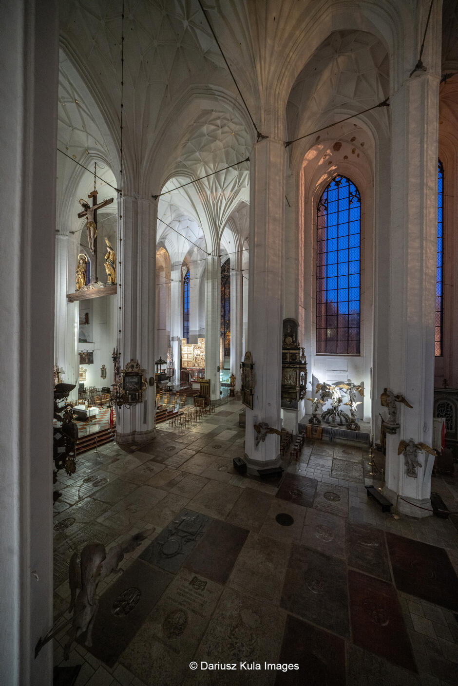 Zdjęcie ukazuje monumentalne wnętrze gotyckiej świątyni, prawdopodobnie katedry lub bazyliki, o majestatycznej i surowej architekturze. Wysokie, strzeliste sklepienia krzyżowo-żebrowe oraz smukłe, białe kolumny nadają wnętrzu lekkości i potęgują wrażenie przestrzeni. W centralnej części obrazu widoczna jest zawieszona belka tęczowa z realistycznymi rzeźbami Chrystusa na krzyżu oraz towarzyszących mu postaci, co jest charakterystycznym elementem wystroju gotyckich kościołów. W tle dostrzec można ołtarz główny, bogato zdobiony i podświetlony, który kontrastuje z surowością kamiennych ścian. Posadzka świątyni pokryta jest płytami nagrobnymi, co wskazuje na historyczne znaczenie tego miejsca oraz tradycję pochówków wewnątrz kościoła. Przy filarach umieszczone są barokowe epitafia i rzeźby aniołów, które dodają wnętrzu sakralnego charakteru.