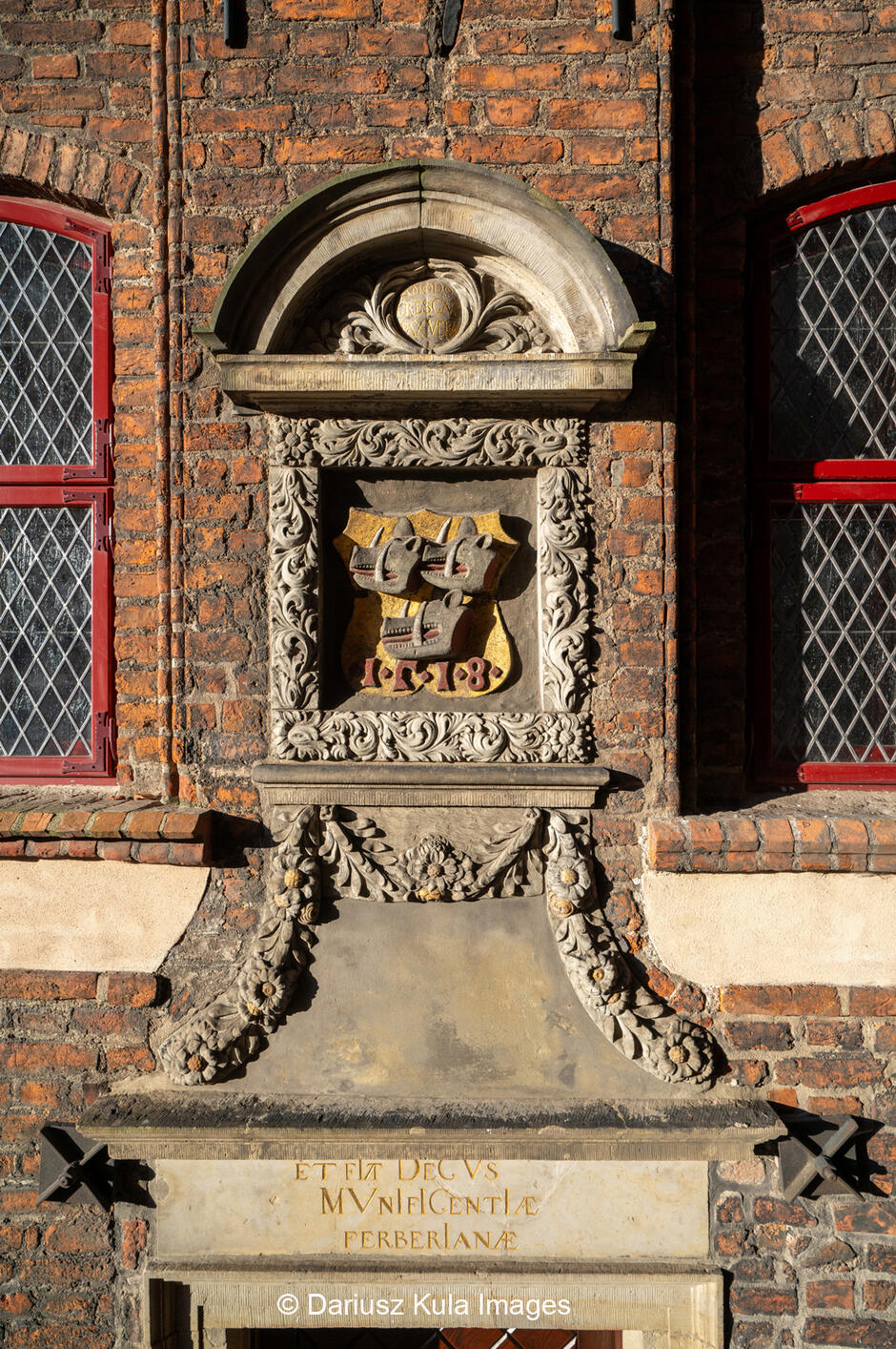 Zdjęcie przedstawia fragment fasady zabytkowego budynku z cegły, ozdobionego bogato rzeźbionymi elementami kamiennymi. Centralnym punktem jest dekoracyjny herb umieszczony w ozdobnej ramie, przedstawiający dwa stylizowane statki z roku 1518 , co może sugerować związek budynku z handlem morskim lub gildia kupiecką. Powyżej herbu znajduje się półokrągła nisza z inskrypcją, otoczona dekoracją z motywami roślinnymi, charakterystyczną dla renesansowej lub manierystycznej stylistyki. Poniżej herbu umieszczony jest marmurowy panel z łacińskim napisem:  ET FIA DECUS MVNIFICENTIAE FERBERIANAE , co można przetłumaczyć jako odniesienie do hojności i zasług rodu Ferberów – wpływowej gdańskiej rodziny patrycjuszy.