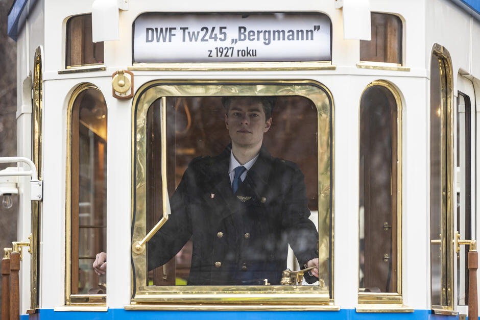 Na zdjęciu widzimy motorniczego ubranego w elegancki, historyczny mundur, prowadzącego zabytkowy tramwaj DWF Tw245 „Bergmann” z 1927 roku. Tramwaj ma stylowe, złote wykończenia i drewniane elementy wnętrza, co podkreśla jego klasyczny charakter