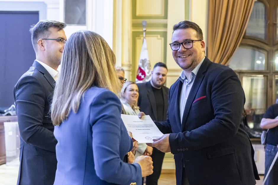 na zdjęciu kobieta w długich włosach wręcza dokument mężczyźnie w garniturze