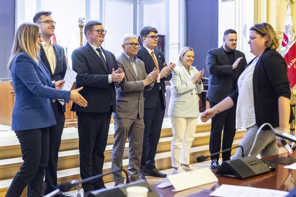 na zdjęciu stoi w rzędzie siedmioro radnych w oficjalnych strojach, biją brawo