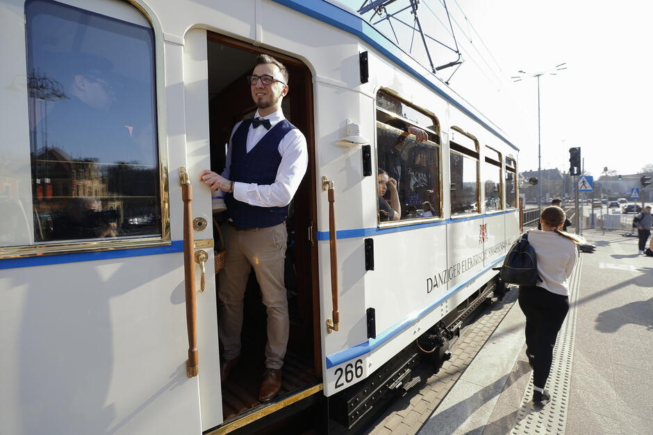 Stary tramwaj w kolorze kremowym, z którego drzwi wychyla się na przystanku elegancko ubrany konduktor