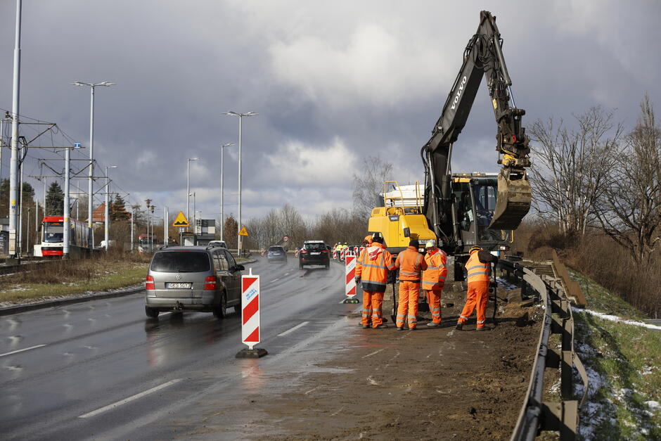 na zdjęciu drogowy w pomarańczowych kombinezonach, stoją przy żółtej koparce na ulicy, obok jadą samochody
