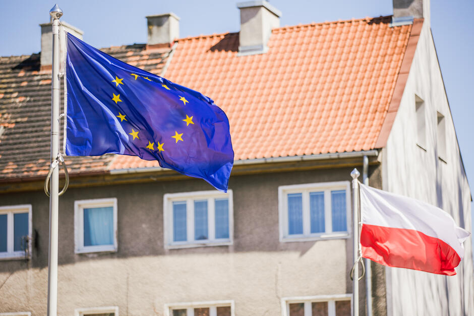 Na zdjęciu widoczne są dwie powiewające na wietrze flagi: Unii Europejskiej po lewej stronie oraz Polski po prawej. W tle znajdują się budynki z czerwonymi dachami i szarymi elewacjami, co sugeruje miejskie otoczenie.