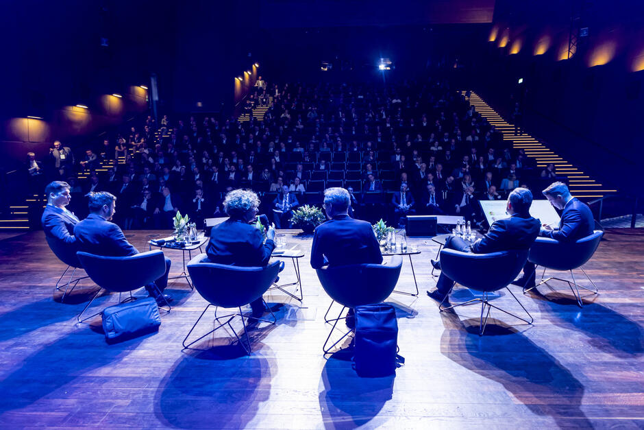 Na zdjęciu widzimy panel dyskusyjny odbywający się podczas Baltic Nuclear Energy Forum. Sześciu panelistów siedzi w półokręgu na scenie, prowadząc rozmowę na temat związany z energetyką jądrową. Jeden z uczestników trzyma mikrofon, co sugeruje, że wypowiada się w danym momencie. Przed nimi znajdują się stoliki z wodą i dekoracjami. Publiczność, licznie zgromadzona w audytorium, uważnie słucha debaty. W tle widoczne są podświetlone schody i nowoczesne oświetlenie sali konferencyjnej, co nadaje wydarzeniu profesjonalny charakter. Niebieskie oświetlenie sceny i kontrastujące ciepłe światła na ścianach podkreślają formalną, ale dynamiczną atmosferę spotkania.