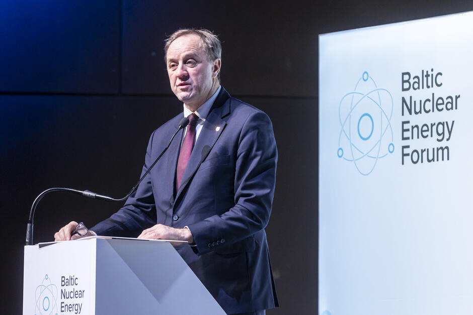 Na zdjęciu widzimy mężczyznę w garniturze i bordowym krawacie, stojącego za podium podczas wystąpienia na Baltic Nuclear Energy Forum. Ma poważny i skupiony wyraz twarzy, a jego postawa sugeruje pewność siebie i zaangażowanie w temat. W jednej ręce trzyma okulary, co może wskazywać, że chwilę wcześniej korzystał z notatek.