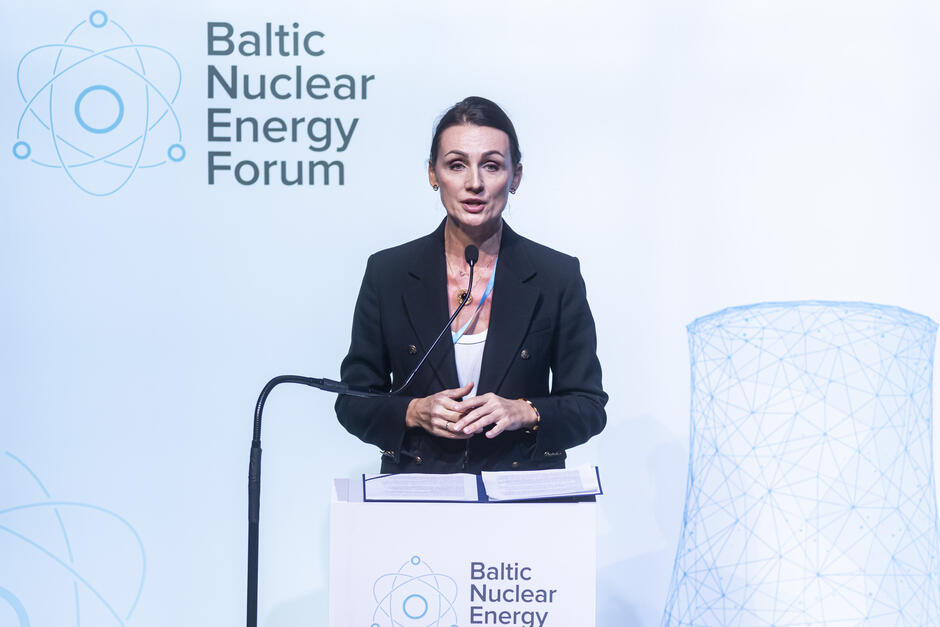 Na zdjęciu widzimy kobietę w eleganckim czarnym żakiecie, stojącą za podium i przemawiającą podczas Baltic Nuclear Energy Forum. Jest profesjonalnie ubrana, a jej postawa i wyraz twarzy sugerują pewność siebie i zaangażowanie w temat. Na podium widoczne są notatki, co wskazuje na dobrze przygotowane wystąpienie.