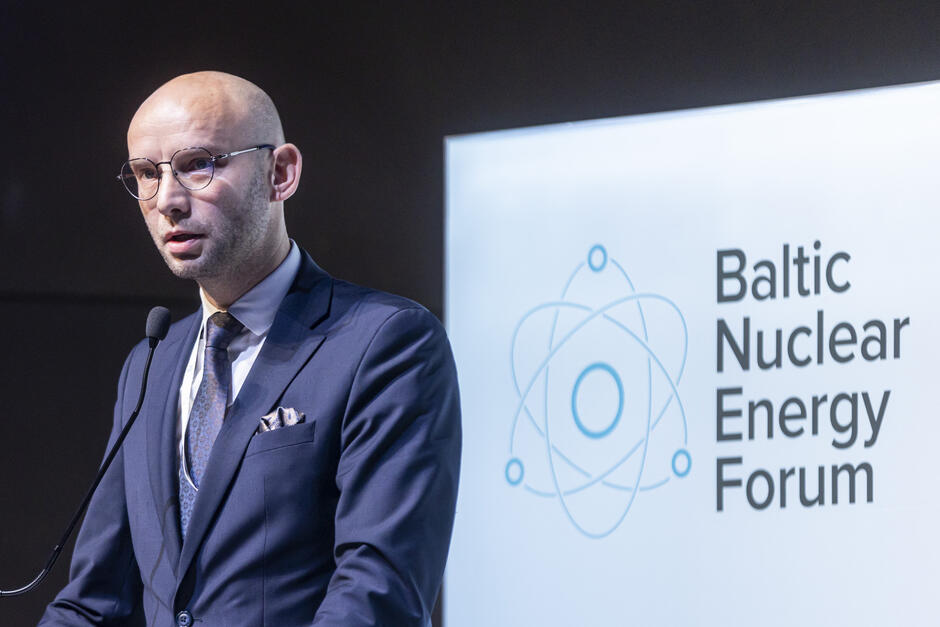 Na zdjęciu widzimy mężczyznę w eleganckim, ciemnym garniturze, który przemawia podczas Baltic Nuclear Energy Forum. Ma na sobie krawat w subtelny wzór oraz ozdobną poszetkę, co podkreśla jego profesjonalny wizerunek. Okulary dodają mu powagi, a skupiony wyraz twarzy sugeruje, że omawia ważne kwestie związane z tematyką wydarzenia.