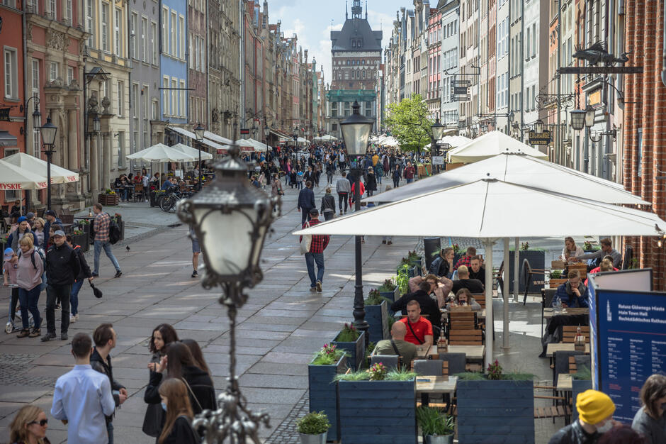 Deptakiem na starym mieście w Gdańsku spacerują wiosennie ubrani ludzie. Część ludzi siedzi przy stolikach w ogródkach restauracji, które ciągną się wzdłuż promenady. Po bokach pierzeje renesansowych kamienic, w tle góruje baszta. 