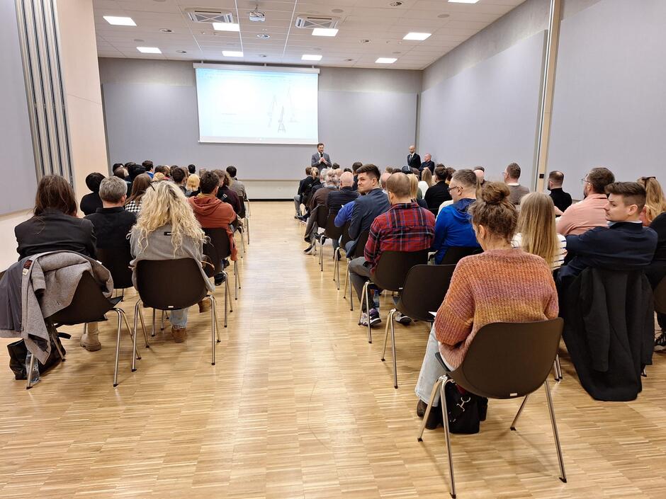 Na sali konferencyjnej siedzą na krzesłach ludzie. Naprzeciw stoi dwóch mężczyzn w garniturach, zwróconych do tłumu twarzami. Za nimi na ścianie wisi ekran.