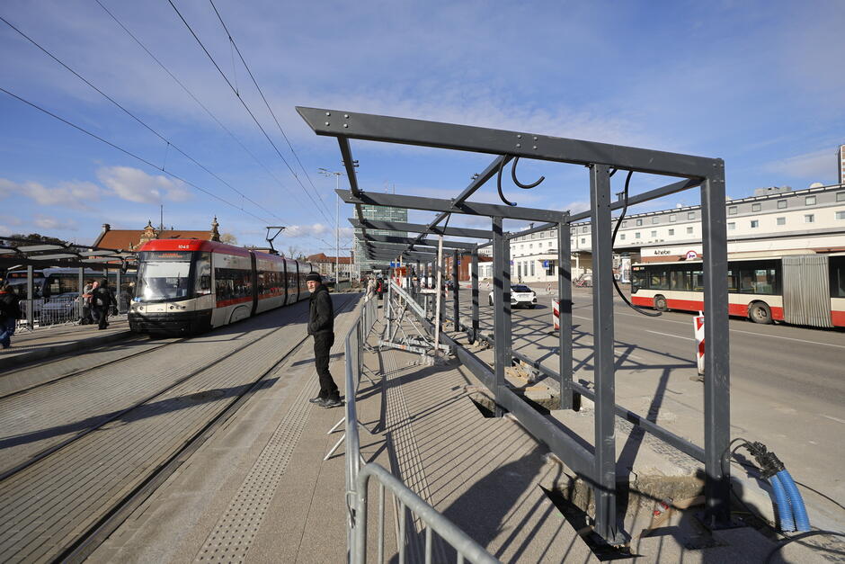 Wiata przystankowa na peronie tramwajowym otoczona barierkami metalowymi, po lewej tor tramwajowy