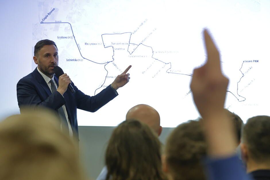 na zdjęciu mężczyzna w średnim wieku, w granatowej marynarce, mówi do mikrofonu, za nim wyświetla się duża prezentacja