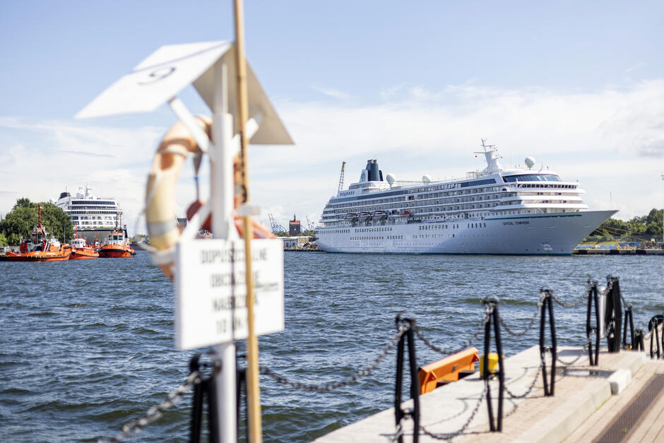 To zdjęcie wykonane z nabrzeża w porcie Gdańsk przedstawia dwa luksusowe statki wycieczkowe – na pierwszym planie Crystal Symphony, a w tle drugi cruiser, którego nazwy nie widać wyraźnie. Kadłub Crystal Symphony lśni w słońcu, a elegancka sylwetka statku przykuwa uwagę na tle portowej infrastruktury.