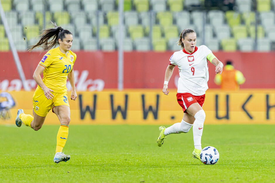 Na zdjęciu widać mecz piłki nożnej kobiet pomiędzy reprezentacjami Polski i Rumunii. Zawodniczka w białym stroju z numerem 9 prowadzi piłkę, podczas gdy przeciwniczka w żółtym stroju z numerem 20 stara się ją dogonić.