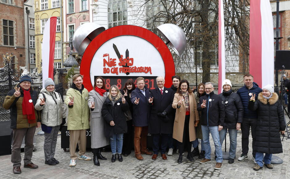 Na zdjęciu widzimy grupę osób stojących na podwyższeniu przed Fontanną Neptuna w Gdańsku, znajdującą się na Długim Targu. Tło stanowią zabytkowe kamienice oraz Dwór Artusa – charakterystyczne budowle w centrum Gdańska. Osoby na scenie ubrane są w zimowe lub jesienne stroje – płaszcze, kurtki, szaliki. Część z nich ma przypięte okrągłe znaczki, co może sugerować udział w jakimś wydarzeniu społecznym lub politycznym. Obok sceny widać biało-czerwone flagi, co wskazuje na polski kontekst uroczystości.
