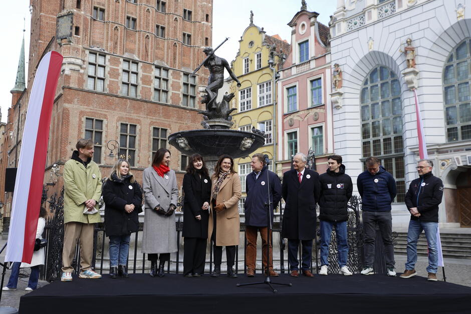 Na zdjęciu widzimy grupę osób stojących na podwyższeniu przed Fontanną Neptuna w Gdańsku, znajdującą się na Długim Targu. Tło stanowią zabytkowe kamienice oraz Dwór Artusa – charakterystyczne budowle w centrum Gdańska. Osoby na scenie ubrane są w zimowe lub jesienne stroje – płaszcze, kurtki, szaliki. Część z nich ma przypięte okrągłe znaczki, co może sugerować udział w jakimś wydarzeniu społecznym lub politycznym. Obok sceny widać biało-czerwone flagi, co wskazuje na polski kontekst uroczystości. W centrum zdjęcia znajduje się mikrofon, co sugeruje, że mogło mieć miejsce przemówienie lub briefing prasowy. Atmosfera wydaje się spokojna i przyjazna.