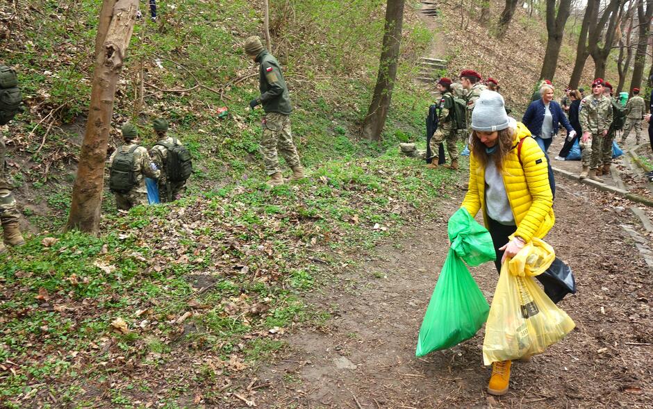 Las. Dziewczyna w żółtej kurtce z dwoma workami wypełnionymi śmieciami. W tle młodzież z liceum mundurowego