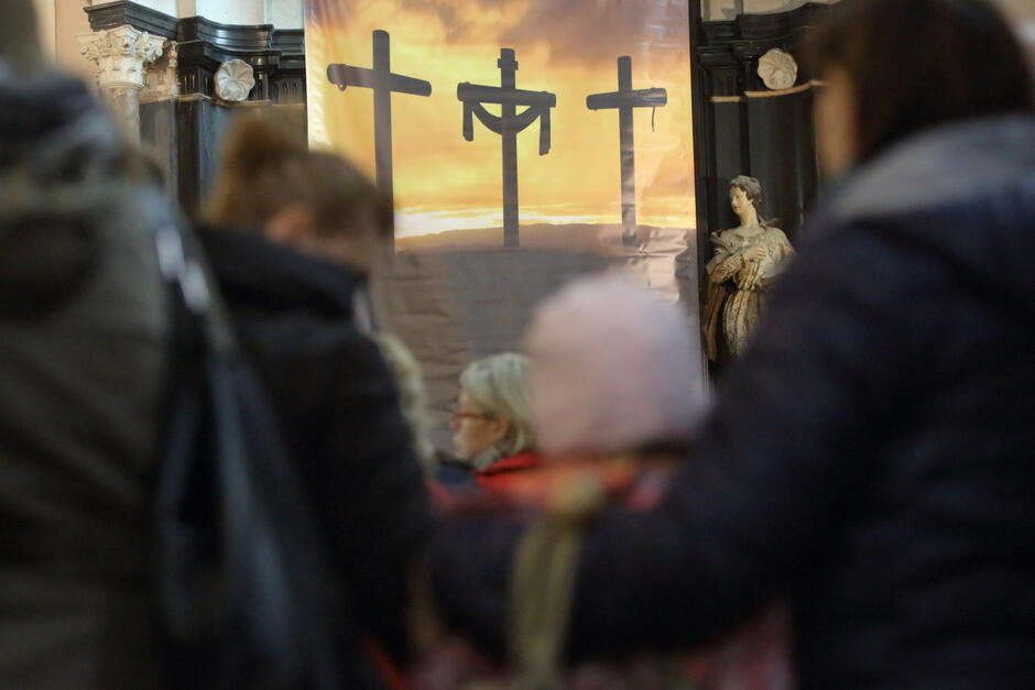 Na zdjęciu widać grupę ludzi wewnątrz kościoła, skierowanych w stronę baneru z trzema krzyżami na tle zachodzącego słońca, symbolizującego ukrzyżowanie.