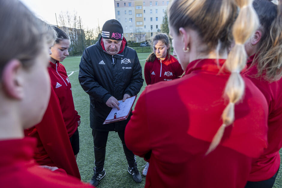 grupa młodych dziewcząt i trener stojący na boisku piłkarskim. Trener nosi czarną kurtkę z czerwono-białym logo po lewej stronie klatki piersiowej. Trzyma schowek i wydaje się coś wyjaśniać dziewczynom. W grupie jest sześć dziewczyn, wszystkie w czerwonych kurtkach, i uważnie patrzą na trenera. W tle widoczne są budynki i drzewa.