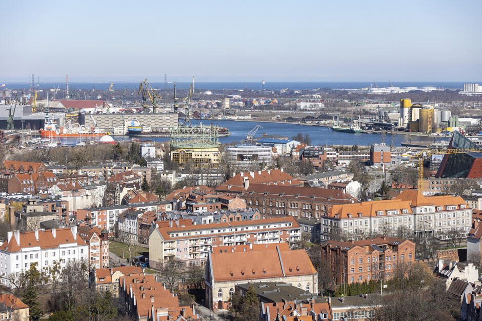 Na zdjęciu widać panoramę Gdańska z charakterystyczną zabudową Starego Miasta, ceglanymi kamienicami i czerwonymi dachami. W tle rozciąga się port morski z licznymi dźwigami i statkami, świadczący o przemysłowym charakterze nadmorskiej części miasta.
