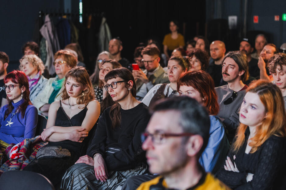 Publiczność słucha uważnie wystąpienia, patrząc w stronę sceny z różnym wyrazem twarzy – od skupienia po lekką zadumę. Sala jest pełna, co świadczy o dużym zainteresowaniu wydarzeniem