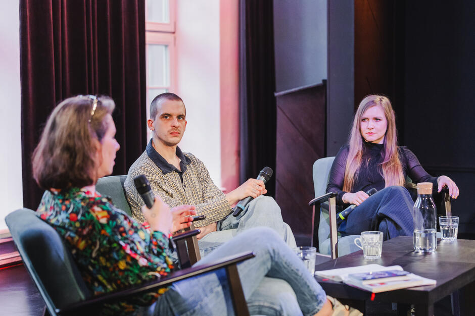 Trzy osoby uczestniczą w panelu dyskusyjnym – każda z mikrofonem w ręce, siedząc w wygodnych fotelach. Atmosfera rozmowy wydaje się poważna i skoncentrowana, a wymiana zdań toczy się w spokojnym tempie