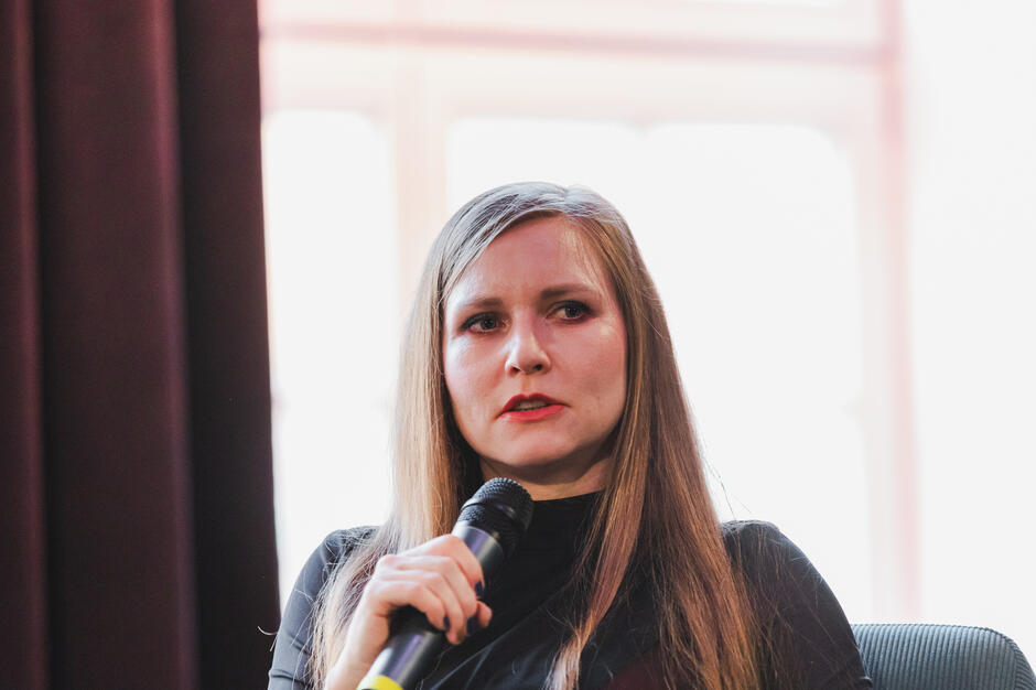 Kobieta z długimi, jasnymi włosami mówi do mikrofonu z wyraźnym zaangażowaniem. Jej mimika wskazuje na intensywność wypowiedzi, co sugeruje, że poruszany temat jest dla niej ważny