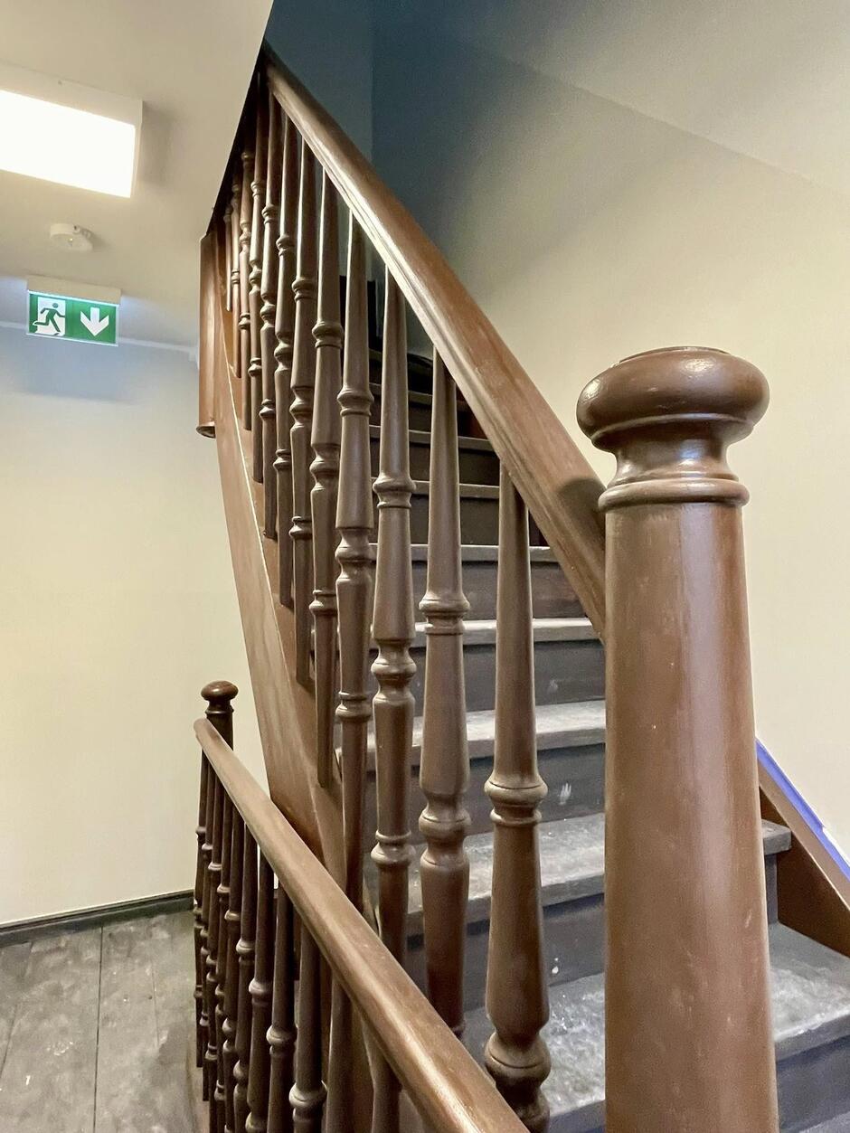 Na zdjęciu widoczne są drewniane schody wewnętrzne z klasyczną balustradą wykonaną z ciemnego drewna. Balustrada składa się z równo rozmieszczonych, ozdobnych tralek i zaokrąglonej poręczy. Drewno ma połysk, choć widać na nim lekkie ślady zużycia i drobne rysy. Schody prowadzą na wyższe piętro budynku. Na górze kadru widoczny jest znak ewakuacyjny z zieloną strzałką skierowaną w dół oraz piktogramem biegnącej osoby, wskazujący kierunek wyjścia ewakuacyjnego. Ściany są w jasnych, neutralnych kolorach, co dobrze kontrastuje z ciemnym drewnem. Całość sprawia wrażenie wnętrza budynku mieszkalnego lub biurowego z elementami klasycznego wystroju.