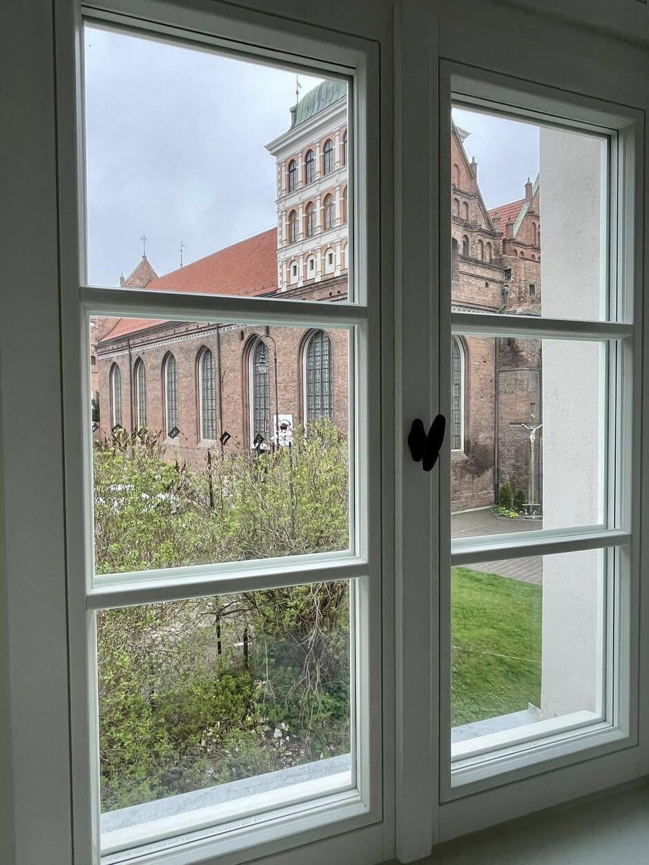 Na zdjęciu widzimy widok z wnętrza budynku przez klasyczne, białe, drewniane okno. Za oknem rozciąga się widok na imponującą, zabytkową architekturę – najprawdopodobniej jest to kościół lub klasztor. Ceglane mury, wysokie ostrołukowe okna oraz wieża z zielonym dachem i białymi detalami architektonicznymi wskazują na styl gotycko-renesansowy. W dolnej części kadru widoczny jest fragment ogrodu – z zieloną trawą i krzewami, które dopiero zaczynają się zazieleniać, co sugeruje wiosenną porę roku. Pogoda za oknem wydaje się pochmurna, niebo jest szare, co jeszcze bardziej podkreśla surowość i majestat historycznego budynku. Cała scena sprawia spokojne, niemal klasztorne wrażenie.