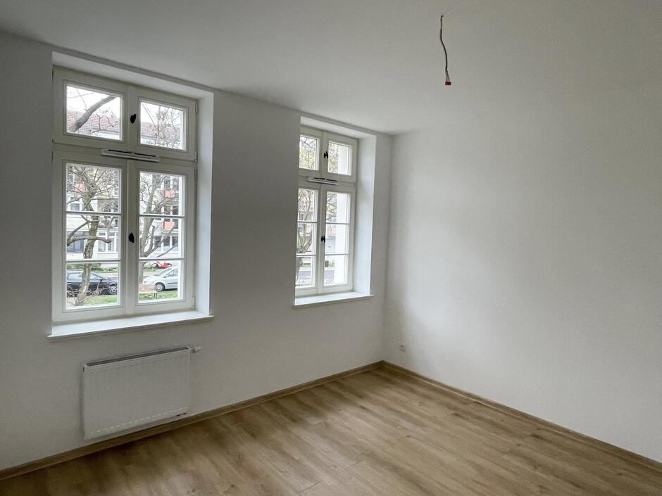 Na zdjęciu widzimy wnętrze jasnego, świeżo wyremontowanego pokoju. Pomieszczenie ma białe ściany i sufit, a na podłodze ułożono panele w naturalnym, jasnym odcieniu drewna. Brakuje jeszcze oświetlenia – z sufitu zwisa przewód elektryczny, co sugeruje, że instalacja elektryczna czeka na zamontowanie lampy. W pokoju znajdują się dwa duże, podwójne okna z białymi ramami, przez które wpada sporo naturalnego światła. Na zewnątrz widać drzewa oraz zaparkowane samochody, co sugeruje, że mieszkanie znajduje się w miejskiej okolicy – prawdopodobnie w budynku zlokalizowanym przy spokojnej ulicy z zabudową mieszkaniową.