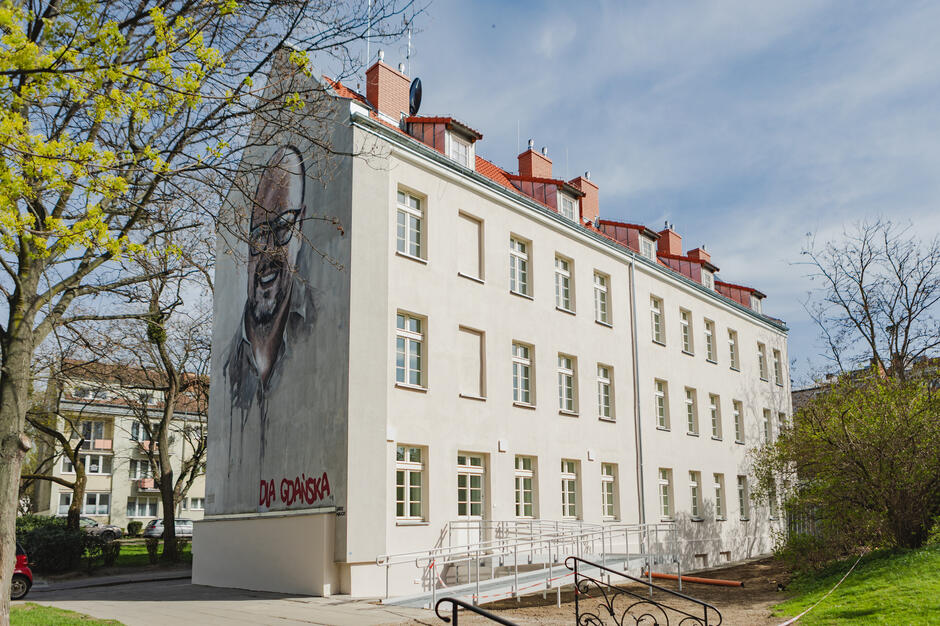 Na zdjęciu widzimy biały, odnowiony budynek o kilku kondygnacjach, z czerwonym dachem i licznymi oknami. Elewacja jest czysta i schludna, a przy wejściu znajduje się podjazd dla osób z niepełnosprawnościami. Po lewej stronie budynku, na bocznej ścianie, znajduje się duży mural przedstawiający uśmiechniętego mężczyznę w okularach. Pod jego wizerunkiem widnieje napis: Dla Gdańska  (po polsku: Dla Gdańska), co sugeruje, że osoba ta miała znaczenie dla miasta Gdańsk. 