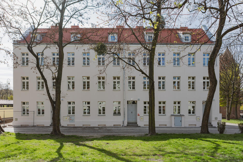 Na zdjęciu widzimy frontową fasadę odnowionego budynku, utrzymaną w jasnej kolorystyce. Budynek ma trzy kondygnacje nadziemne oraz poddasze użytkowe, widoczne dzięki oknom wychodzącym z dachu pokrytego czerwonymi dachówkami. Elewacja jest symetryczna i estetyczna, z dużą liczbą okien rozmieszczonych równomiernie. Pośrodku znajduje się wejście główne z niewielkimi schodkami i drzwiami pomalowanymi na jasno.