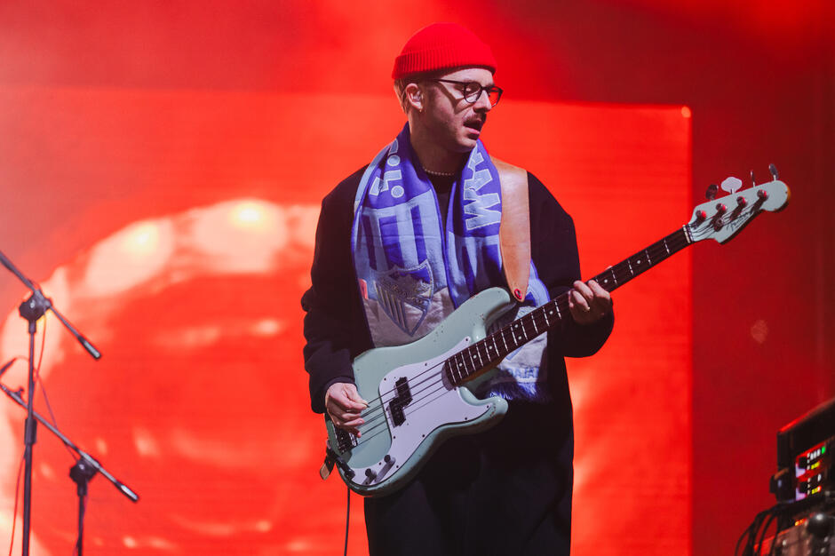 Na zdjęciu widać muzyka grającego na gitarze basowej podczas koncertu, oświetlonego intensywnym czerwonym światłem scenicznym. Artysta ma na sobie czerwono pomarańczową czapkę, okulary, niebieski szalik z herbem piłkarskim i czarny płaszcz