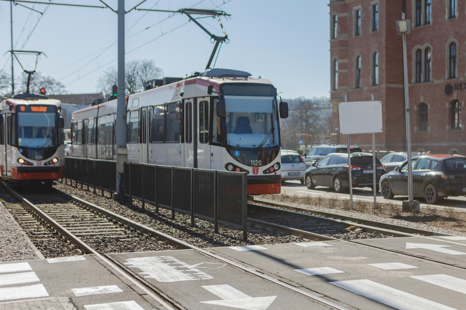 Na zdjęciu widać dwa tramwaje jadące równoległymi torami w centrum miasta, przy przystanku z wyznaczonym przejściem dla pieszych. W tle znajdują się zaparkowane samochody oraz charakterystyczny, ceglany budynek, co sugeruje miejskie otoczenie o zabytkowym charakterze.