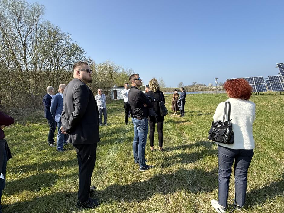 Zdjęcie przedstawia grupę osób podczas wizyty terenowej przy instalacji paneli fotowoltaicznych na otwartym, zielonym terenie w słoneczny dzień.