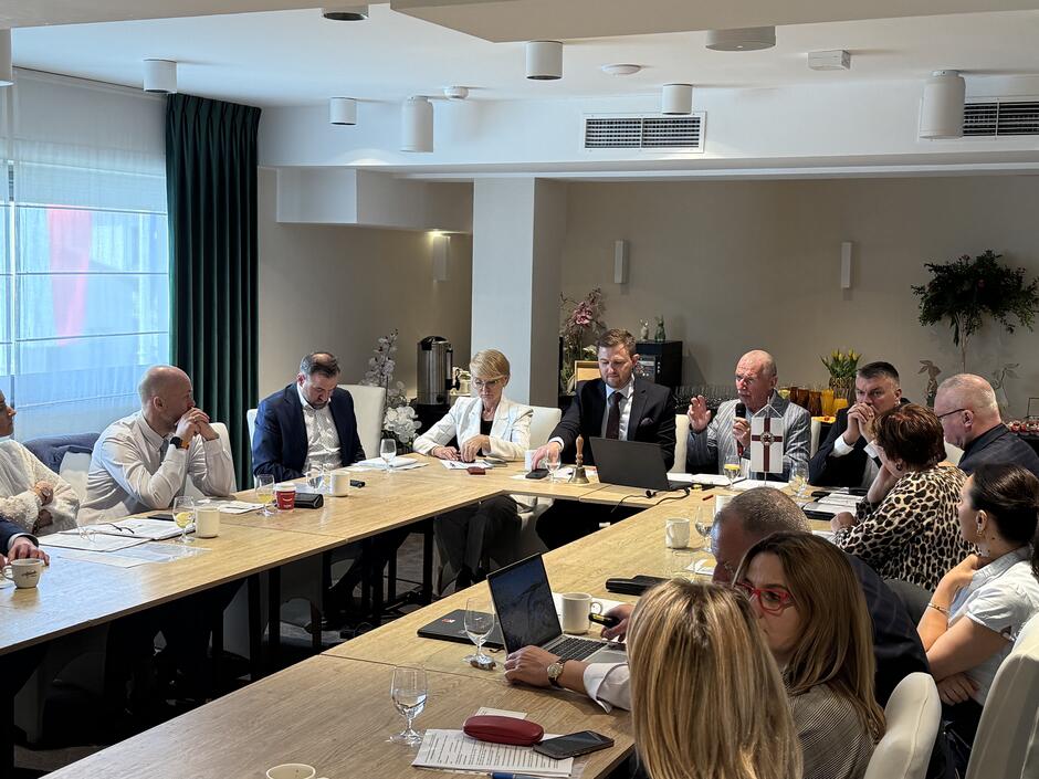 Zdjęcie przedstawia spotkanie biznesowe lub konferencję, podczas której grupa osób siedzi przy stole, omawiając dokumenty i pracując na laptopach.