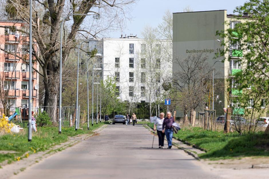 na zdjęciu asfaltowa jezdnia, idą po niej dwie osoby, w tle po obu stronach ulicy widać fragmenty bloków mieszkalnych