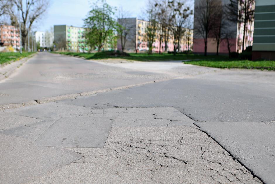 na zdjęciu zniszczona nawierzchnia jezdni, widać liczne spękania, w tle kilka bloków mieszkalnych