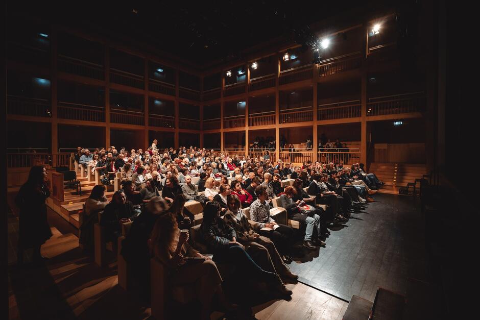 Przedstawia pełną widownię w nowoczesnym, drewnianym wnętrzu teatru lub sali wykładowej. Ludzie siedzący na widowni wyglądają na skupionych i zainteresowanych, czekając na rozpoczęcie wydarzenia lub słuchając wystąpienia