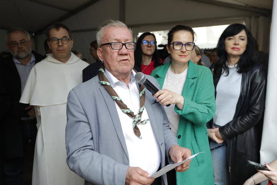 Kilka osób, mężczyzna stojoący w środku grupy, mówi przez mikrofon
