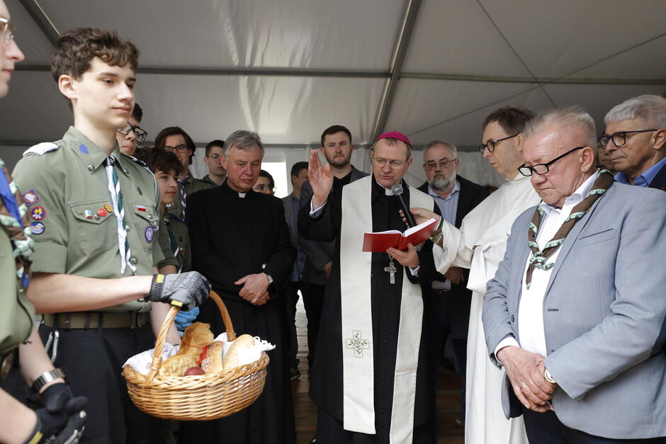 Biskup z kropidłem, ustawiony twarzą do koszyka z pokarmami, który jest trzymany przez harcerza, stojącego po lewej. Wokół biskupa tłumek gości