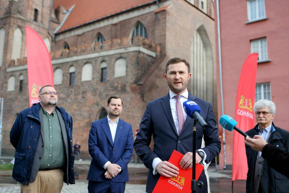 Na zdjęciu widać konferencję prasową odbywającą się na tle historycznego budynku w Gdańsku – mężczyzna w garniturze przemawia do mediów, trzymając teczkę z herbem miasta. W tle stoją inni uczestnicy wydarzenia oraz dwie czerwone flagi z napisem „GDAŃSK”, co wskazuje na oficjalny charakter spotkania.