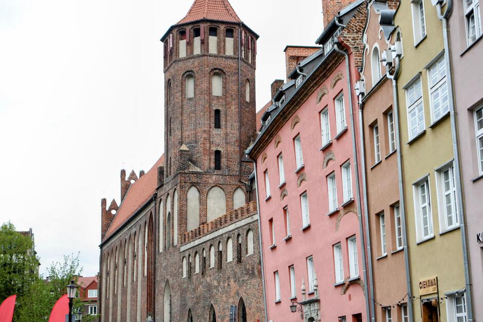 Na zdjęciu widoczna jest urokliwa ulica w Gdańsku z zabytkowym kościołem z czerwonej cegły, który dominuje nad kolorowymi kamienicami o pastelowych elewacjach. Charakterystyczna architektura oraz obecność lokalu „Ciekawa Café” nadają miejscu wyjątkowy, historyczno-kawiarniany klimat.