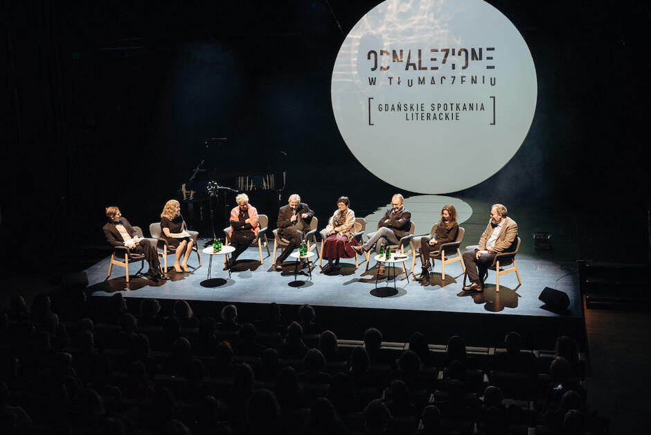Na zdjęciu widzimy panel dyskusyjny zorganizowany na scenie podczas wydarzenia literackiego Odnalezione w tłumaczeniu – Gdańskie Spotkania Literackie, na którym bierze udział dziewięć osób siedzących w półokręgu. W tle znajduje się duży ekran z nazwą wydarzenia, a całość odbywa się przy przytłumionym świetle sceny, co nadaje spotkaniu kameralny, refleksyjny charakter