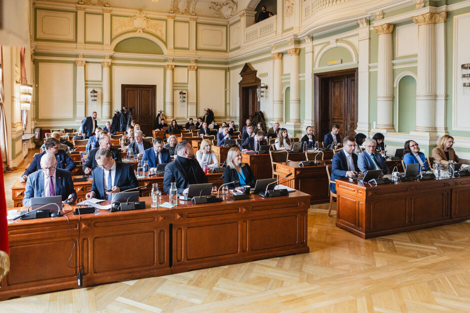 Zdjęcie przedstawia sesję rady miejskiej odbywającą się w eleganckiej sali z klasycystycznym wystrojem i drewnianymi ławami. Uczestnicy, ubrani formalnie, skupiają się na dokumentach i laptopach, co świadczy o powadze i oficjalnym charakterze spotkania.