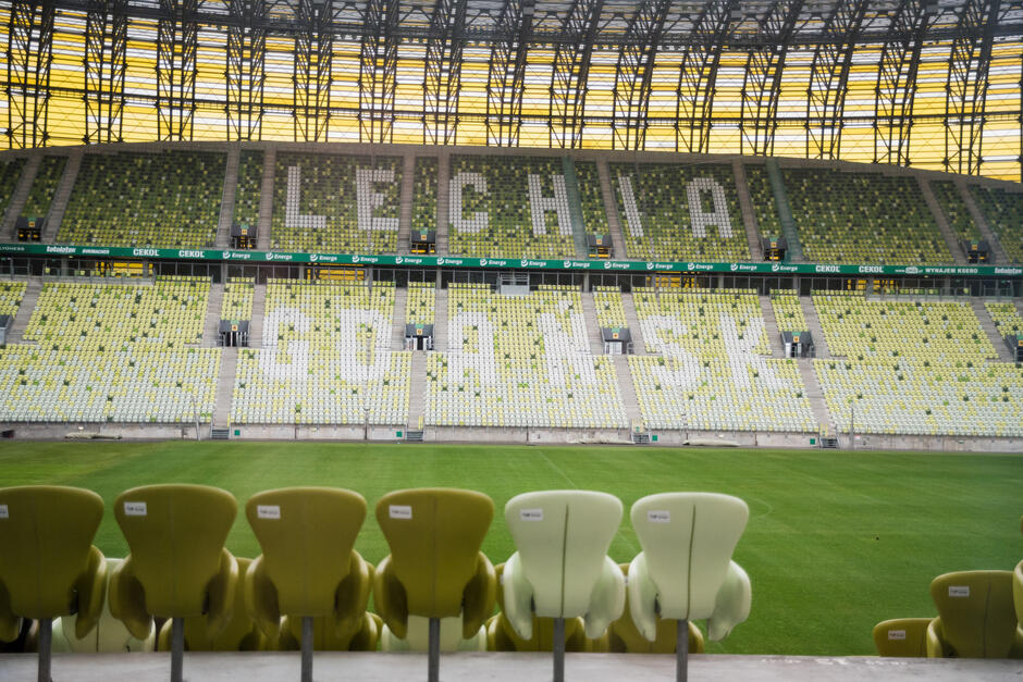 Widok stadionu z rzędami żółtych krzeseł na stadionie. Krzesła są ustawione w rzędach zwrócone na lewą stronę, a na pierwszym planie znajduje się duże zielone pole z trawą. Po prawej stronie tego pola znajduje się kilka żółtych krzeseł, które mają przed sobą czarne metalowe słupy. W lewym górnym rogu znajduje się słowo LECHIA dużymi białymi literami na brązowym tle.